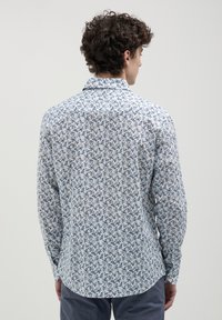 Uomo con capelli ricci che indossa una camicia bianca a maniche lunghe con un piccolo motivo floreale blu, visto di spalle su uno sfondo neutro.