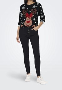 Svart stickad tröja med ett renmotiv som har horn och ett röd band, tillsammans med högmidjade svarta jeans och vita sneakers.