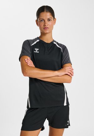 Junge Frau mit dunklem Haar in schwarzer und grauer Sportbekleidung, die die Arme verschränkt hat und vor einem schlichten weißen Hintergrund steht.