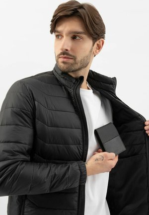 Giacca a vento nera con colletto alto, caratterizzata da un design a righe orizzontali. Una portafoglio elegante in mano, presentato su una maglietta bianca.