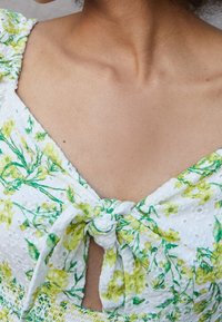 Tessuto bianco con motivo floreale giallo e verde, caratterizzato da un design a scollo a V e un accento con nodo frontale all'collo. Superficie testurizzata.