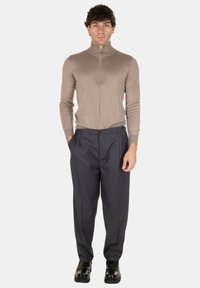 Maglione beige con zip realizzato in tessuto morbido, abbinato a pantaloni grigi scuri a pieghe. Modello che indossa scarpe nere, in piedi contro uno sfondo bianco.