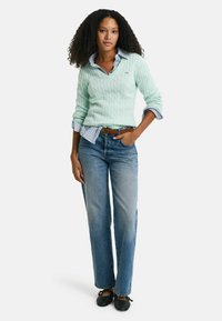 Maglione a trecce verde chiaro indossato sopra una camicia a righe blu, abbinato a jeans dritti azzurri e scarpe nere senza lacci.