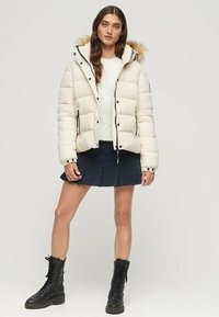 Kremowa kurtka puchowa z futrzanym kapturem, zapinana na zamek z czarnymi akcentami, w połączeniu z granatową plisowaną spódnicą i czarnymi bojowymi butami.