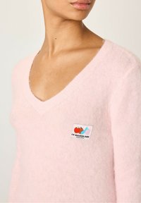 Pull en V rose, fabriqué dans un tissu doux et texturé, avec un écusson logo brodé représentant des illustrations de fruits sur le côté gauche.