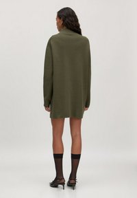 Übergroßes olivgrünes Strickkleid mit langen Ärmeln, gerader Schnitt und gerippten Akzenten am Saum. Das Modell trägt schwarze, durchsichtige Socken und Absätze.