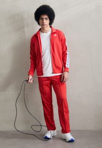 Survêtement rouge en tissu lisse, comprenant une veste à zip intégral et un pantalon assorti avec des rayures sur les côtés. Le modèle tient une corde à sauter noire.
