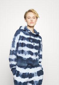 Hoodie em tie-dye azul e branco, fabricado em tecido suave, com capuz com cordão, bolsa frontal e cintura elástica. Design simples e casual.