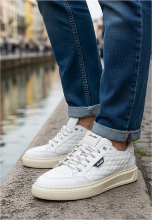 White quilted sneakers with beige soles worn with rolled-up blue jeans standing on a stone ledge by a canal.