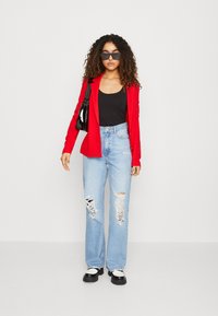 Red blazer, black tank top, distressed light blue jeans, and black platform shoes. Accessories include black sunglasses and a shiny handbag.
