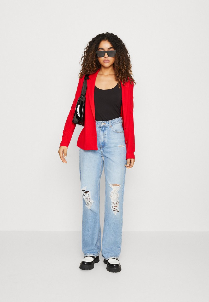 Red blazer, black tank top, distressed light blue jeans, and black platform shoes. Accessories include black sunglasses and a shiny handbag.