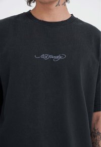Sort bomulds T-shirt med frynset krave, prydet med en lyseblå broderet "Ed Hardy" logo på brystet. Korte ærmer, afslappet pasform.