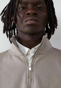 Beige Kapuzenjacke mit hohem Kragen und silbernem Reißverschluss, über einem weiß geringelten Hemd getragen. Glatte Textur mit genähten Details.