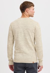 Beige neulepaita, jossa on teksturoitu kuvio, pyöreä kaula-aukko sekä resorit hihansuissa ja helmassa. Mukana on pieni brändilogo alas selässä.