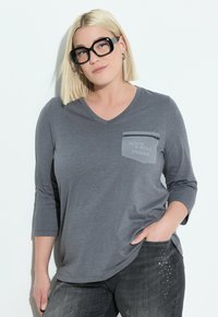 Vrouw met een blonde bob-kapsel, draagt zwarte oversized bril, een grijze V-hals t-shirt met een 'Rock & Roll Inside' zak, en donkere jeans met versieringen.