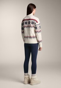 Krém színű, kötött pulóver piros és tengerészkék Fair Isle mintákkal, tengerészkék leggingssel és bézs színű, szőrmebéléssel ellátott csizmával párosítva.