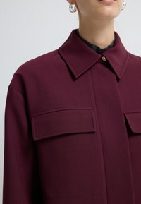 Bordeaux jasje met een gestructureerde kraag, twee voorzakken en een enkele gouden knoopsluiting, gemaakt van een textuurstof.