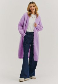 Fialový, nadýchaný dlhý sveter s otvoreným predným dielom a rebrovanými manžetami, v kombinácii s tmavomodrými zavarenými džínsami a svetlosivými nazúvacími topánkami.