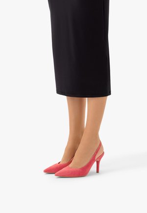 Jambes portant une jupe noire et des talons hauts à bout pointu en daim corail rose, à bride arrière avec un talon fin, sur fond blanc.