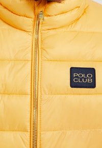 Gul puffjacka med quiltad textur, med en guldfärgad dragkedja och en kvadratisk marinblåklapp med texten "POLO CLUB" på bröstet.