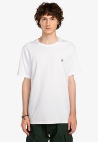 Hvid bomulds T-shirt med rund halsudskæring, korte ærmer og et lille sort emblem på brystet. Glat tekstur, afslappet pasform.