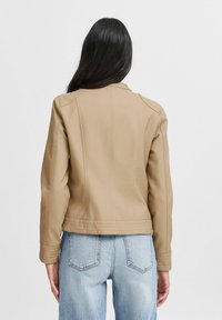 Chaqueta de piel sintética beige con diseño ajustado, costuras verticales y cuello acanalado. Combinada con jeans de tiro alto de color azul claro.