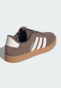 Smeđe nubuk tenisice s bijelim trostrukim prugama, gumeni potplat u boji gume, okrugli vrh i teksturirane vezice, s Adidas logotipom sa strane.