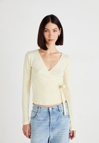 Lysgul, ribbet langærmet wrap-top med bindebånd i taljen og en dyb V-hals. Parret med lyse denimjeans.