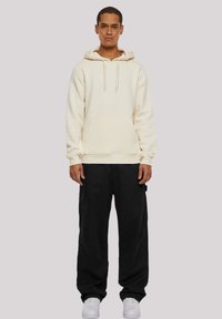 Cream-colored hoodie with a kangaroo pocket, drawstring hood, ribbed cuffs. Paired with loose black pants and white sneakers.