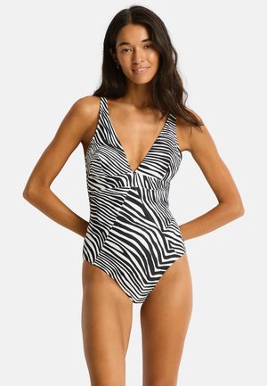 Kobieta z długimi ciemnymi włosami w czarno-białym jednoczęściowym kostiumie kąpielowym w zebrę, stojąca z rękami za plecami.