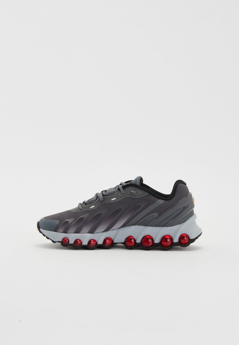 Zapatilla deportiva gris con parte superior de malla transpirable, suela curvada y esferas de amortiguación rojas. Presenta sutiles patrones en forma de onda y puntera reforzada.