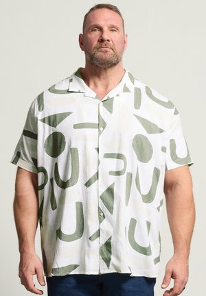 Mann mit kurzem Bart, der ein weißes kurzärmeliges Hemd mit abstrakten grünen Formen vor einem einfarbigen hellen Hintergrund trägt.