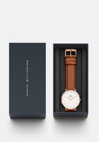 Zegarek Daniel Wellington w granatowym pudełku. Posiada różowo-złotą kopertę, białą tarczę oraz brązowy skórzany pasek z zapięciem.