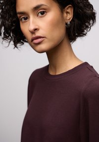 Bordowa koszula z długim rękawem i okrągłym dekoltem, o miękkiej teksturze. Posiada subtelne detale na ramionach oraz gładki, dopasowany krój.