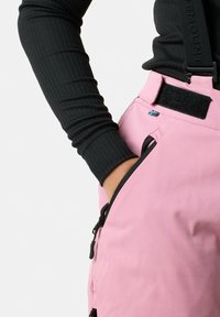 Pink snow pants with adjustable straps, black zippers, and a pocket. The fabric is smooth with a durable finish. Features a small flag accent.