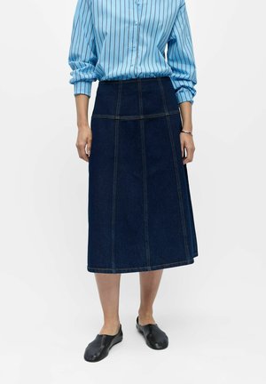 Osoba nosi modrú pruhovanú košeľu s dlhým rukávom, tmavú rifľovú midi sukňu s švami, čierne slip-on topánky a strieborný náramok.