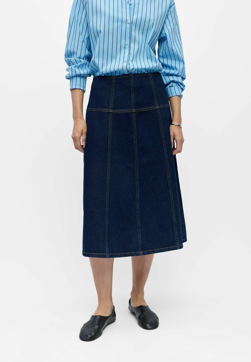 Personne portant une chemise rayée bleue à manches longues, une jupe midi en denim foncé avec des détails de couture, des chaussures noires sans lacets et un bracelet argenté.