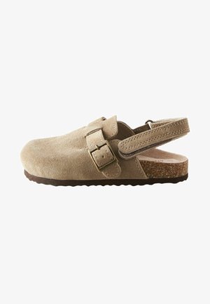 Sandale en suède beige avec une sangle à boucle, dos ouvert, et semelle en caoutchouc texturée, présentant une zone de orteil lisse et une semelle intérieure en liège.