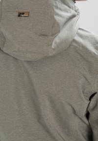 Veste grise à capuche en tissu texturé, dotée d'une petite étiquette de marque sur le haut du dos et de détails de couture visibles.