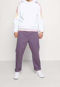 Man met een witte hoodie met oranje en roze strepen op de mouwen, paarse opgedraaide broek en witte sneakers, staand tegen een witte achtergrond.