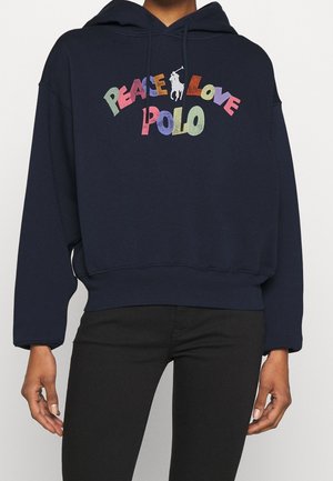 Sweat à capuche bleu marine avec texte multicolore "Peace Love Polo" et petit logo blanc de joueur de polo sur la poitrine, porté avec un pantalon noir.