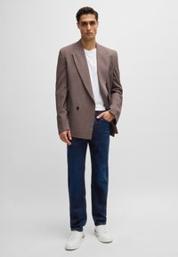 Uomo che indossa un blazer oversize marrone, una maglietta bianca, jeans blu scuro e sneakers bianche, in piedi contro uno sfondo chiaro e semplice.