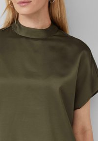 Haut vert olive avec un col montant, des manches courtes et une texture lisse et brillante. Présente des lignes épurées et peu de détails de couture.