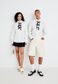 Hvite skjorter med svarte sløyfeaksenter, kombinert med en svart skjørt og beige shorts, og en glinsende sølvveske og fottøy.