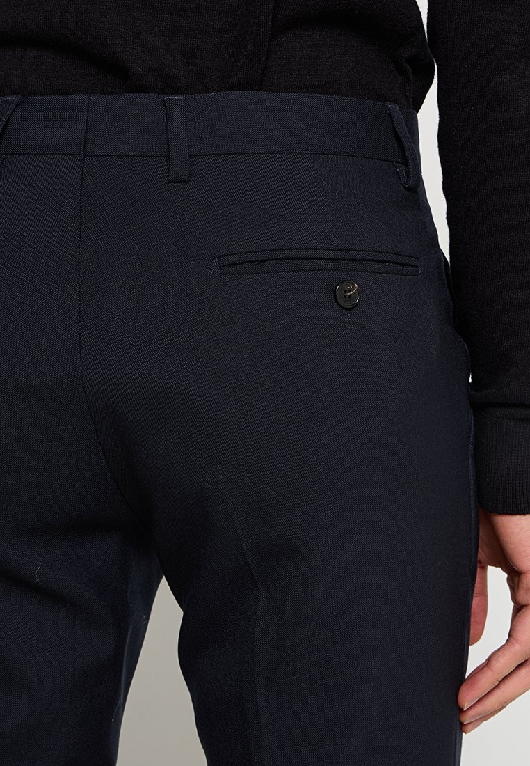 Los pantalones navy presentan un diseño ajustado con una textura suave. Se puede ver un bolsillo trasero con cierre de botón, junto con pasadores para el cinturón.