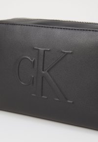 Svart läderväska med präglad "CK"-logotyp, slank rektangulär form, slät yta och silverfärgad dragkedja upptill.