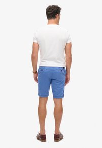 T-shirt bianca da uomo con maniche corte, abbinata a pantaloni corti blu chiaro con tasche posteriori e bottoni a contrasto. Indossata con mocassini marroni.