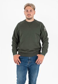 Felpa verde oliva con scollatura rotonda, polsini e orlo a coste, con un patch logo sulla spalla sinistra. Indossata con jeans blu.