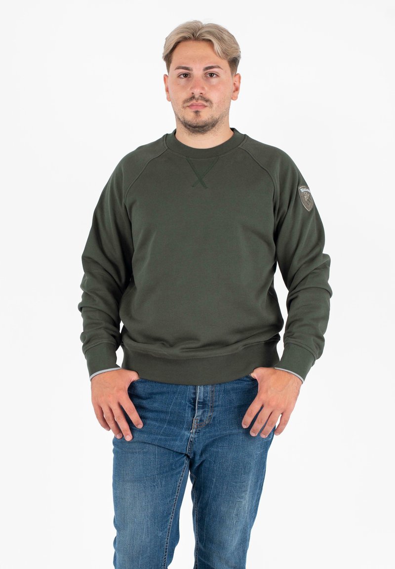 Felpa verde oliva con scollatura rotonda, polsini e orlo a coste, con un patch logo sulla spalla sinistra. Indossata con jeans blu.