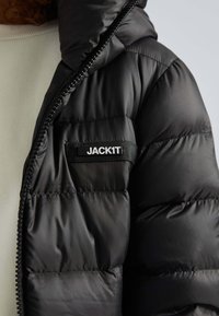 Sort oppustet jakke med quiltet design, der har en lynlås lukning og et mærket skilt med teksten "JACK1T." Glat, skinnende tekstur.